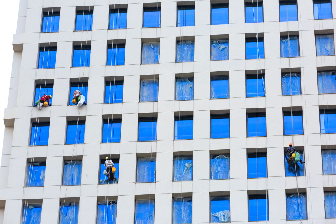 Professional workers clean skyscraper windows using ropes in urban setting.