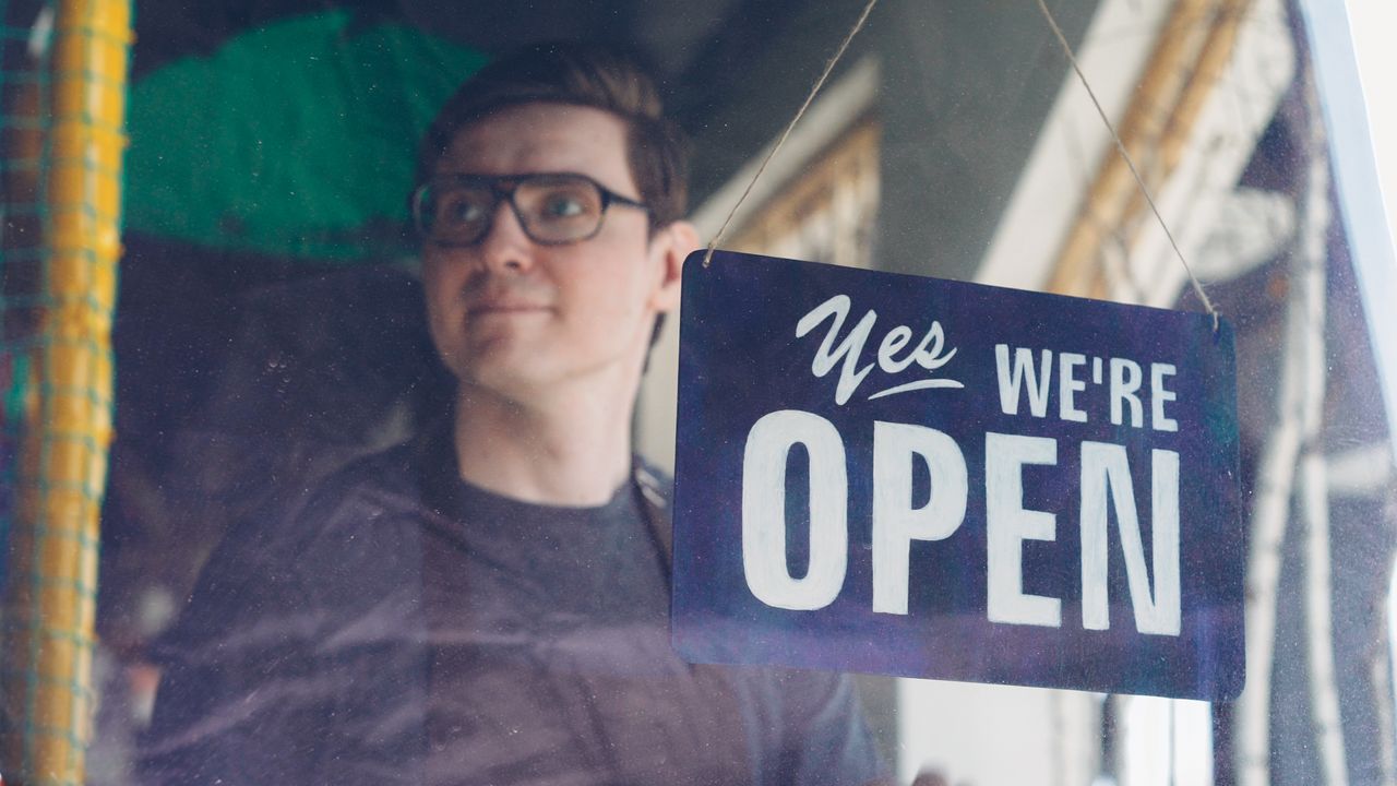 Smiling adult man behind a glass with a 'Yes, We're Open' sign, suggesting a welcoming store.